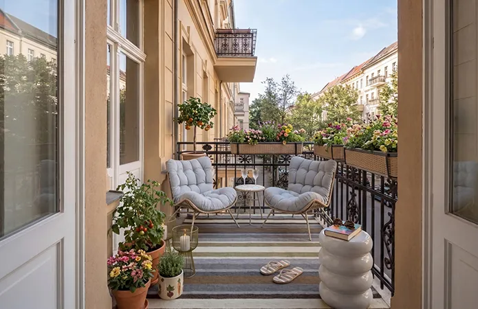 Balkon mit schwarzem Geländer und langen Rattan-Blumenkörben, gestreifter Teppich in Beige-Braun-Blautönen, Loungesessel-Set Bambus mit hellblauen Auflagen und kleinem Tisch, weißer runder Beistelltisch und grünes Windlicht