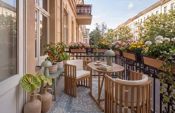 Balkon mit schwarzem Geländer und Balkonmöbel-Set aus Holz mit zwei runden Stühlen und Holzstäben, ovaler passender Tisch, LED-Tischleuchte in mint, Vintage Teppich mit orientalischem Muster, Sitzbank in mint, darauf beige Übertöpfe mit Tomaten und Kräutern, Geländer voll mit länglichen Blumenkästen und bunten Blumen
