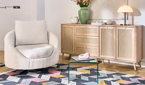 Beiger Sessel, Kommode aus hellem Holz und Rattan Optik, Glas-Beistelltisch, dazu Musterteppich in Dunkelgrau, Gelb, hellblau und Rosa