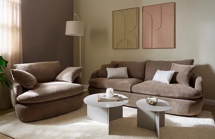Big Sofa und passender größerer Polstersessel in Taupe mit weißem Teppich und zwei runden grauen Couchtischen, zwei Kunstbilder an beiger Wand