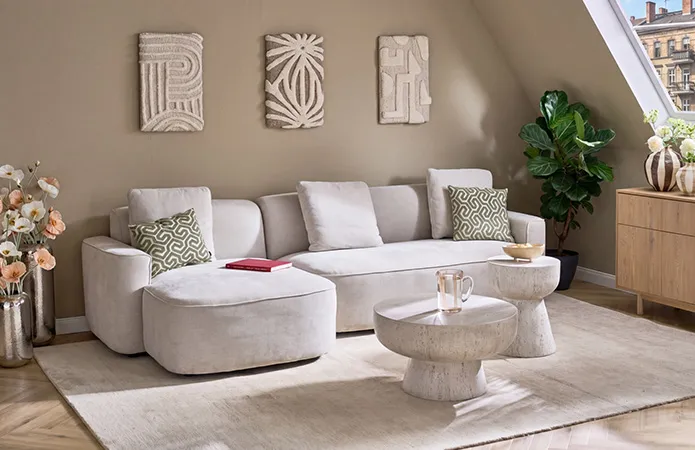 Beiges Ecksofa mit runden Ecken, großer weißer Teppich, runder Couchtisch und passender Beistelltisch in Steinoptik, Stoff-Leinwandbilder an Wand mit verschiedenen Mustern, zwei große Chrom-Bodenvasen mit Pflanzen