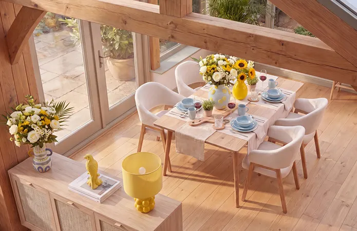 Großer Wintergarten mit Esstisch aus hellem Holz und weißen Stühlen mit Holzfüßen, hellblaues Geschirr, große Blumensträuße mit gelben und hellblauen Vasen, helle Holzkommode in Rattanoptik mit gelber Tischlampe und gelben Dekovogel