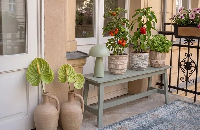 Sitzbank in mintgrün mit braun-gemusterten Übertöpfen und Kräutern, LED-Tischleuchte in Mint, daneben auf dem Boden braune Keramikkrüge mit Pflanzen