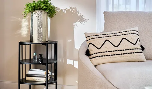 Beiges Kissen mit schwarzen geraden Linien und einer Zickzacklinie auf beigem Sessel, schwarzer Beistelltisch in rund mit Chromvase