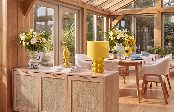 Großer Wintergarten mit Esstisch aus hellem Holz und weißen Stühlen mit Holzfüßen, hellblaues Geschirr, große Blumensträuße mit gelben und hellblauen Vasen, helle Holzkommode in Rattanoptik mit gelber Tischlampe und gelben Dekovogel