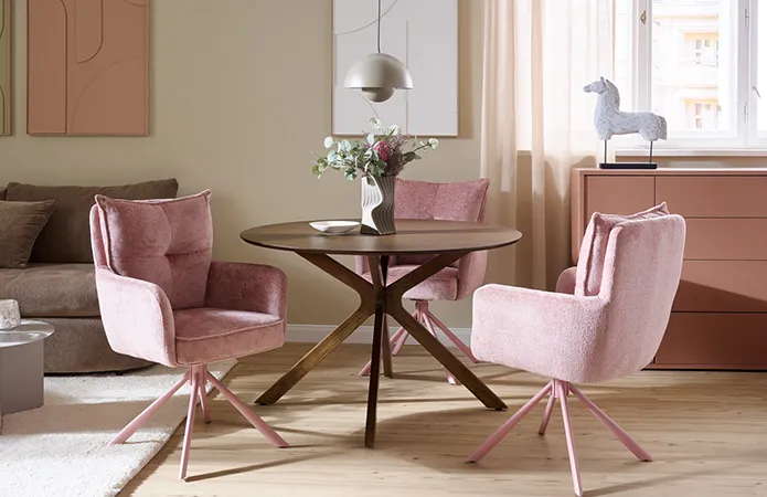 Runder Esstisch aus dunklem Holz mit vier rosanen Polsterstühlen und rosa Beinen, dahinter rostrote Kommode, braungraue 3D Vase auf Tisch mit Sträuchern