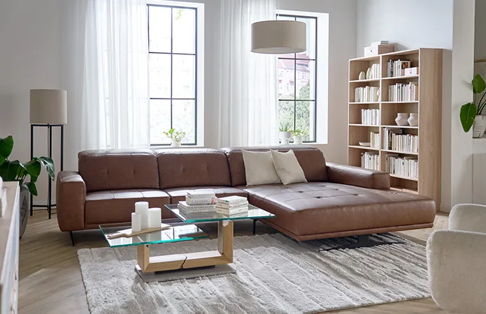 Gradliniges braunes Leder-Ecksofa, flauschiger beiger Teppich, Couchtisch mit dickem Holzfuß und zwei Glasplatten, hohes Holzregal mit Büchern