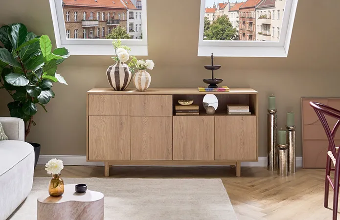 Frontalansicht helles Sideboard unter Dachschräge, drei große Boden-Kerzenständer in Chrom und grünen Kerzen, auf Sideboard beige und braungestreifte Vasen und dunkle Etagére