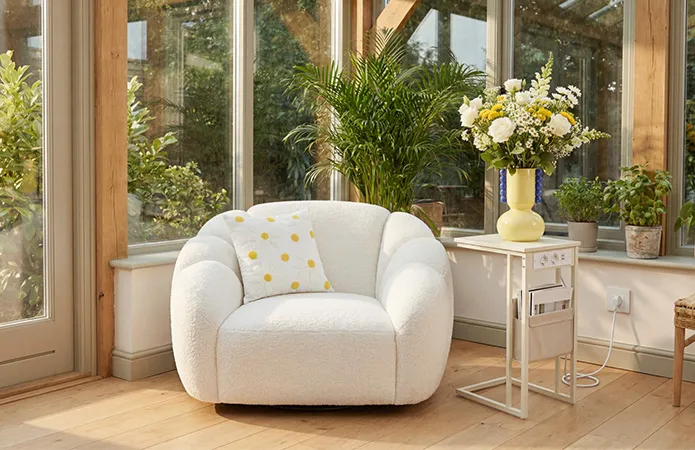 Weißer, runder Sessel mit Boucléstoff, daneben kleiner Beistelltisch in beige mit integrierten Stromsteckern, weißes Dekokissen mit Gänseblümchen und gelben Punkten, gelbe Vase mit Blumenstrauß