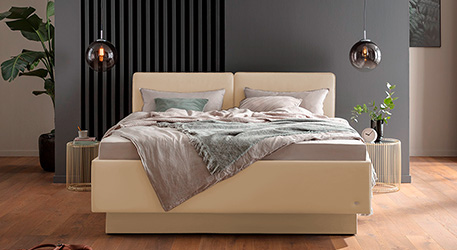 cremefarbiges Polsterbett mit Bettwäsche in Beige, grüner Decke, grünen Kissen, Nachttischen aus Metall, Pendelleuchten vor grauer Wand