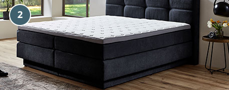 Blaue Markierung 2, dunkelgraues Boxspringbett mit weißem Topper, im Hintergrund Schlafzimmer