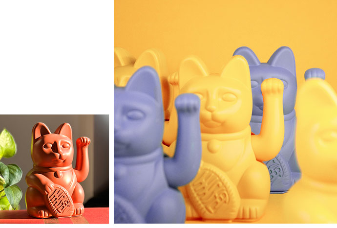 Bild 1: matt orange Winkekatze, Grünpflanze. Bild 2: 2 matte lilane und 3 matte gelbe Winkekatzen vor gelbem Hintergrund