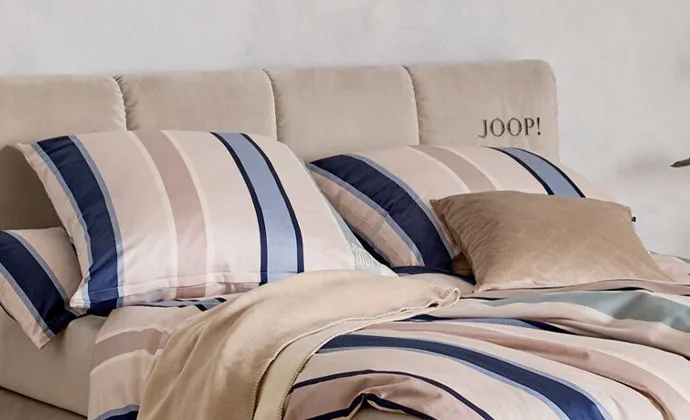eiges Bett mit Joop-Schriftzug und mit Bettwäsche im blau-beigen Streifenmuster, beige Decke und beiges Kissen
