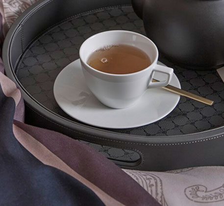 dunkelgraues Tablett mit weißer Tasse mit Tee und Untertasse, Löffel und Textilien in Blau und Lilatönen