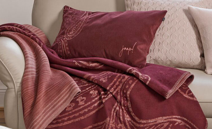 Beiges Ledersofa mit rotem Kissen und roter Decke mit Paisley-Muster und JOOP! Schriftzug, cremefarbigen Kissen mit und ohne Muster und JOOP! Schriftzug