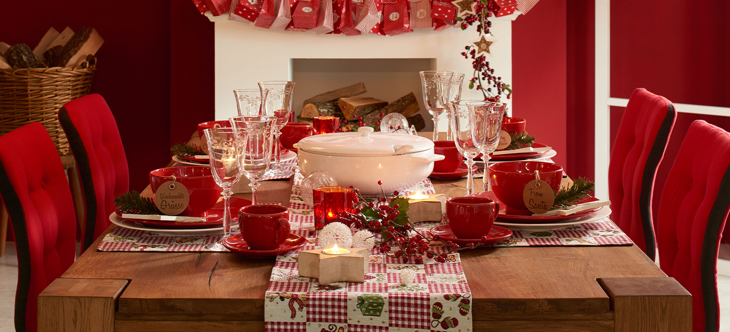 Weihnachtlich gedeckter Holztisch mit rotem Geschirr und Weihnachtsdeko - rote Polsterstühle - im Hintergrund ein Kamin