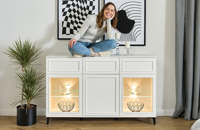 Weißes Sideboards im Landhausstil mit beleuchteten Fächern mit Glasfronten davor. Frau sitzt auf Sideboard