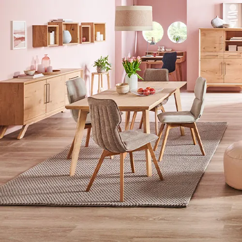 Wohnzimmer im Skandi-Stil mit Holzmöbeln, bestehend aus Sideboard, Wandboard, Esstisch mit grauen Polsterstühlen mit Holzgestell, cremefarbigen Sessel mit Holzgestell, Couchtishc beigem Ecksofa mit Kissen und Plaid, beigen Pouf, Schreibtisch, Highboard, Vitrine, Lowboard, Wandregal, Regal, Textilien, Teppiche, Deko.