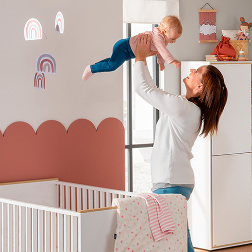 Frau hält Baby in die Luft, helles Babyzimmer mit weißen Möbeln