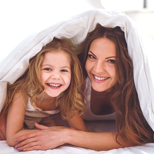 Mädchen und Frau die unter einer weißen Bettdecke liegen, wobei diese auch deren Köpfe bedeckt