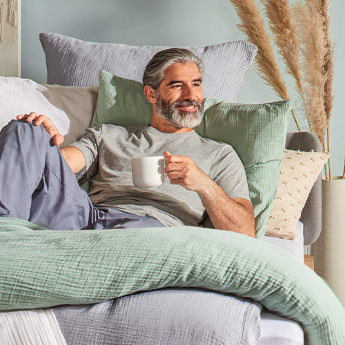 Im Bett liegender Mann mit Kaffeetasse - Bettwäsche aus Musselin in Grün, weiß und Grau  - Vase mit Trockengras