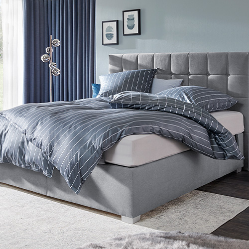 Schlafzimmer mit grauem Boxspringbett mit blaugrau-weiß gestreifter Bettwäsche