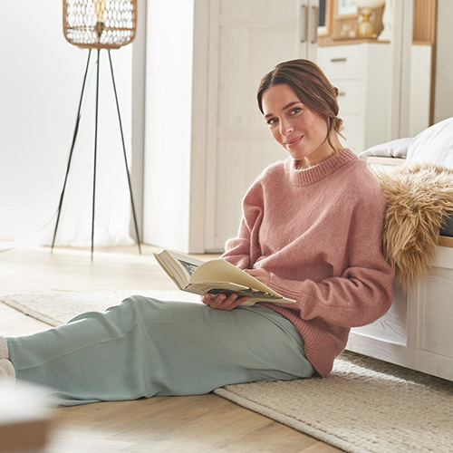 Frau sitzt mit Buch auf dem Teppich vor dem Bett