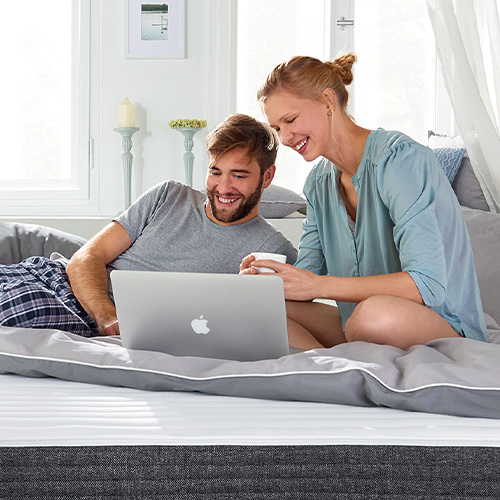 Auf Bett sitzendes Paar, das auf einen Computer schaut - große Matratze in Weiß und Grau - graue Bettwäsche