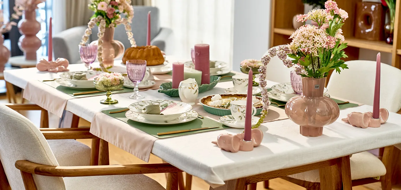 Langer Tisch mit weißer Tischdecke, cremefarbenen Essstühlen und Holzgestell, runde Vasen in Rosa mit Blüten und Pflanzen in weiß und rosa, florales Geschirr in weiß mit grünen Ranken, rosa Schleifen Kerzenständern, grüne Eissschalen, Zitronentarte, Kerzen in mint und rosa, mintfarbene Platzsets