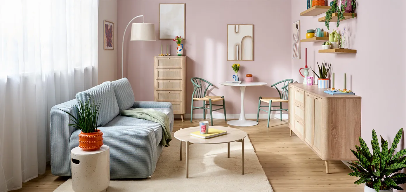 Kleines Wohnzimmer mit hellrosa Wänden, hellen Holzkommoden in Rattanoptik, hellblaues Cord-Sofa als Zweisitzer, beiger Teppich, runder Couchtisch in Kaschmirfarben, kleine Essecke mit rundem Esstisch und zwei Rattanstühlen mit grünem Gestell, bunte Deko aus Übertöpfen, Vasen und Kerzenständern auf Kommoden und Wandregalen