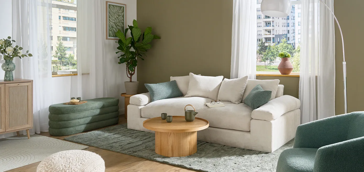 Wohnzimmer mit weißer und olivegrüner Wand, cremefarbenes Sofa als Zweisitzer mit grünen Kissen, daneben eine Polstersitzbank in Grün mit Stauraum, runter Couchtisch aus hellem Holz, grüner Teppich, weiße Bogenleuchte und grüner Sessel aus Boucléstoff
