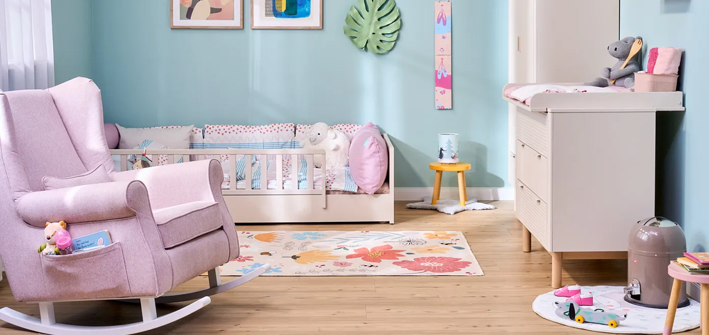 Buntes und verspieltes Kinderzimmer mit hellblauer Wand, Teppich mit bunten Blumen, rosafarbener Schaukelsessel und Seitentasche für Spielzeug, Bodenbett mit Rausfallschutz in Beige, Kommode mit Wickelaufsatz in Beige