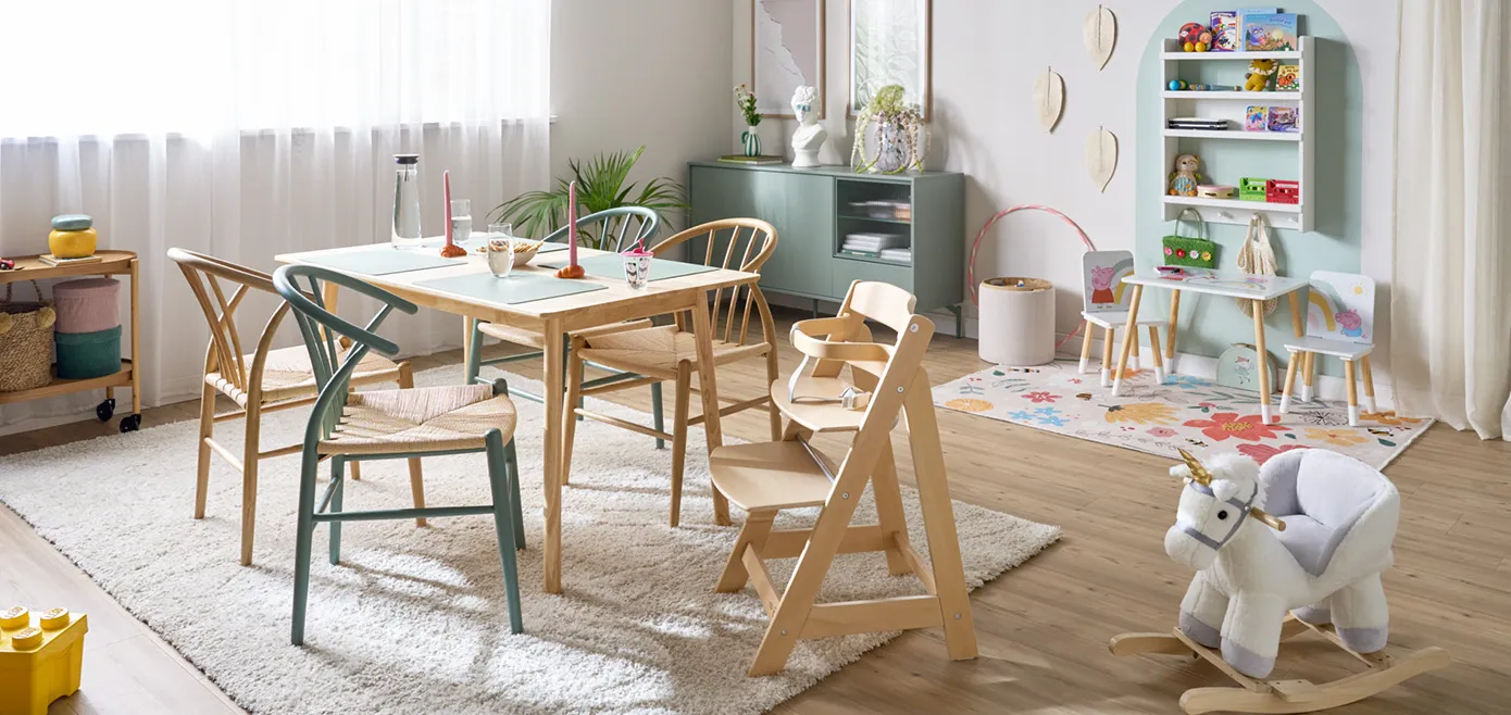 Wonzimmer im Skandistil mit Holz-Esstisch und passenden Holzstühlen, Hochstuhl, Schaukelpferd, grüngraue Kommode, Spielecke mit kleinem Tisch und Stuhlset mit Pepper Pig, kleiner Teppich mit Blumen, Wandregal mit Spielzeug, farblicher Rundbogen an Wand zur Abgrenzung