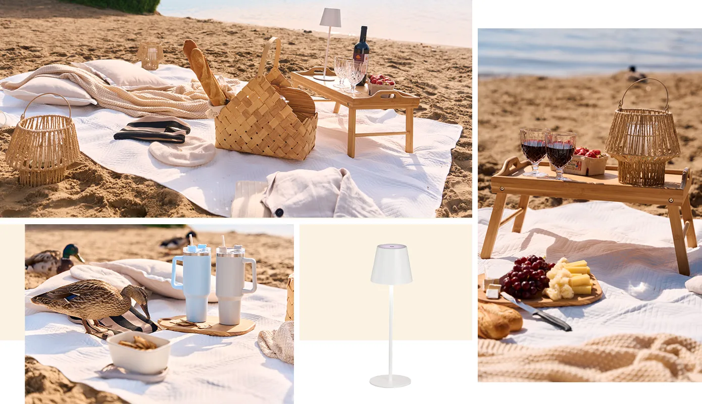 Picknick am Strand mit beiger Tagesdecke, geflochtener Tragekorb mit Brotdosen, Schneidebrett mit Käse und Weintrauben und Baguette, Serviertablett mit Füßen mit Windlicht, zwei Weingläsern und Schale mit Erdbeeren, Stanley Cubs