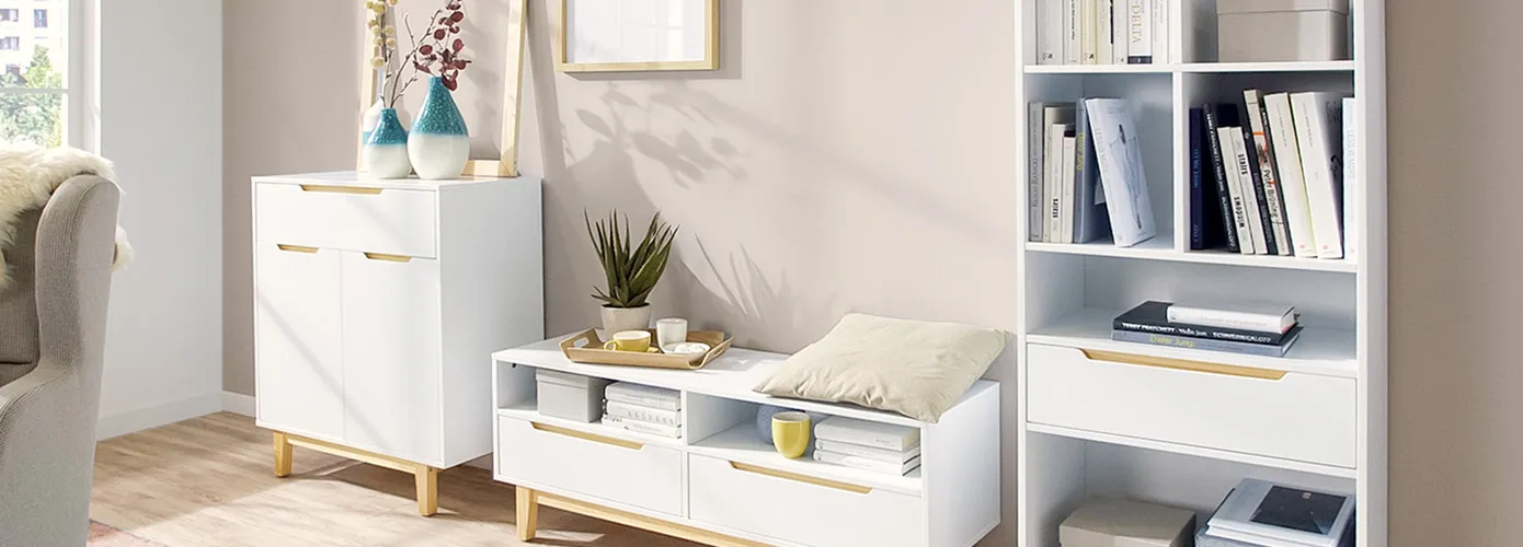 Heller Wohnraum mit Möbeln der Serie Brescia in Weiß und hellem Holz. Kommode, Lowboard und hohes Standregal