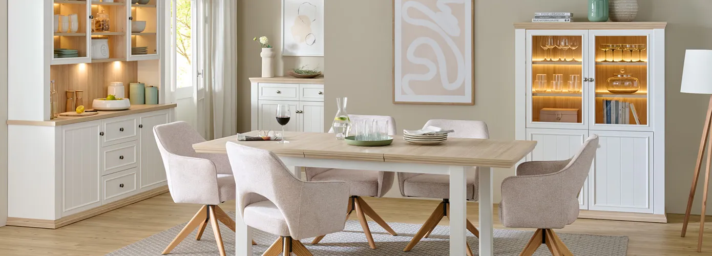 Esszimmer mit weißen Landhausmöbeln mit Holzakzenten, bestehend aus Buffet, Kommode, Vitrine, Esstisch und cremefarbigen Polsterstühlen, Deko