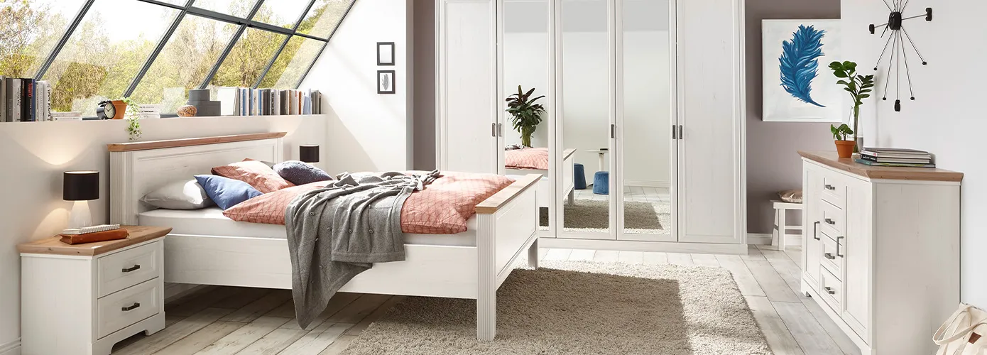 Schlafzimmer mit weißen Landhausmöbeln mit Holzakzenten, bestehend aus Bett mit Nachtkommode, Schrank, Kommode, Textilien und Deko.