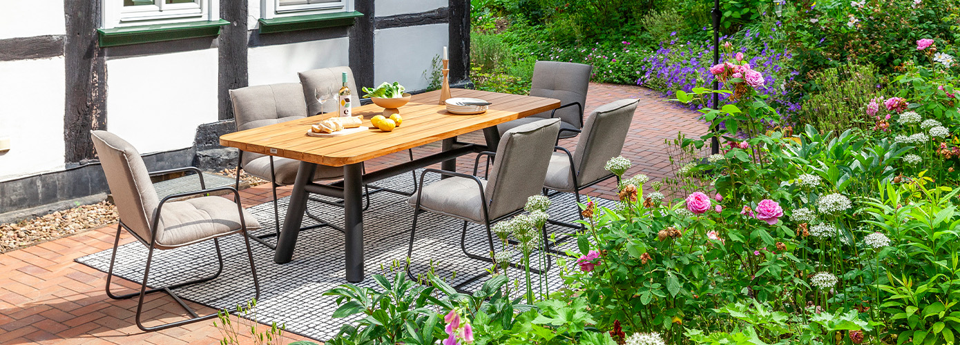Outdoor-Sitzgruppe vor Fachwerkhaus auf Terrasse im Grünen, Esstisch mit Holzplatte und schwarzem Gestell, dazu 6 Polsterstühle mit schlankem, schwarzen Metallgestell
