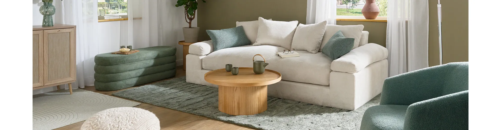 Wohnzimmer mit weißer und olivegrüner Wand, cremefarbenes Sofa als Zweisitzer mit grünen Kissen, daneben eine Polstersitzbank in Grün mit Stauraum, runter Couchtisch aus hellem Holz, grüner Teppich, weiße Bogenleuchte und grüner Sessel aus Boucléstoff