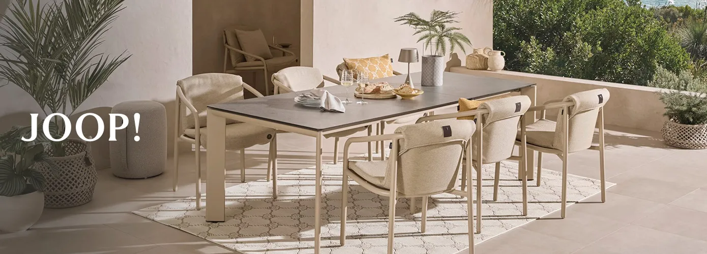 Joop! gedeckter Gartentisch in Beige und dunkelgrauer Tischplatte. Beige Gartenstühle mit beigen Auflagen. Gelbes Kissen mit weißem Muster, weißer Outdoor-Teppich mit beigem Muster auf einer Terrasse